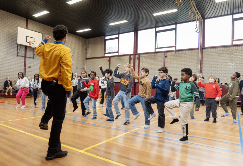 In de gymzaal krijgen kinderen van basisschool De Stromen een dansles. Je ziet de leraar op zijn rug. Verschillende kinderen doen het dansje mee en kijken blij naar de leraar.