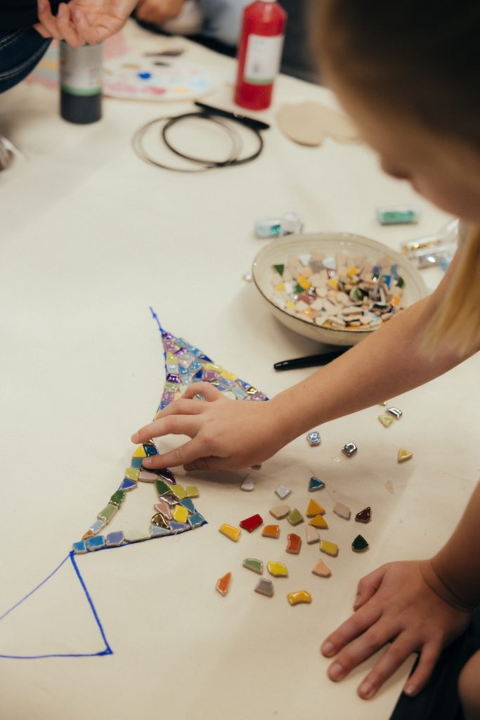 Een ingezoomde foto van het creatieve doek. Je ziet een kinderhand die aan het knutselen is. Het kind maakt een vrolijke vlaggetjeslijn met allerlei kleuren.