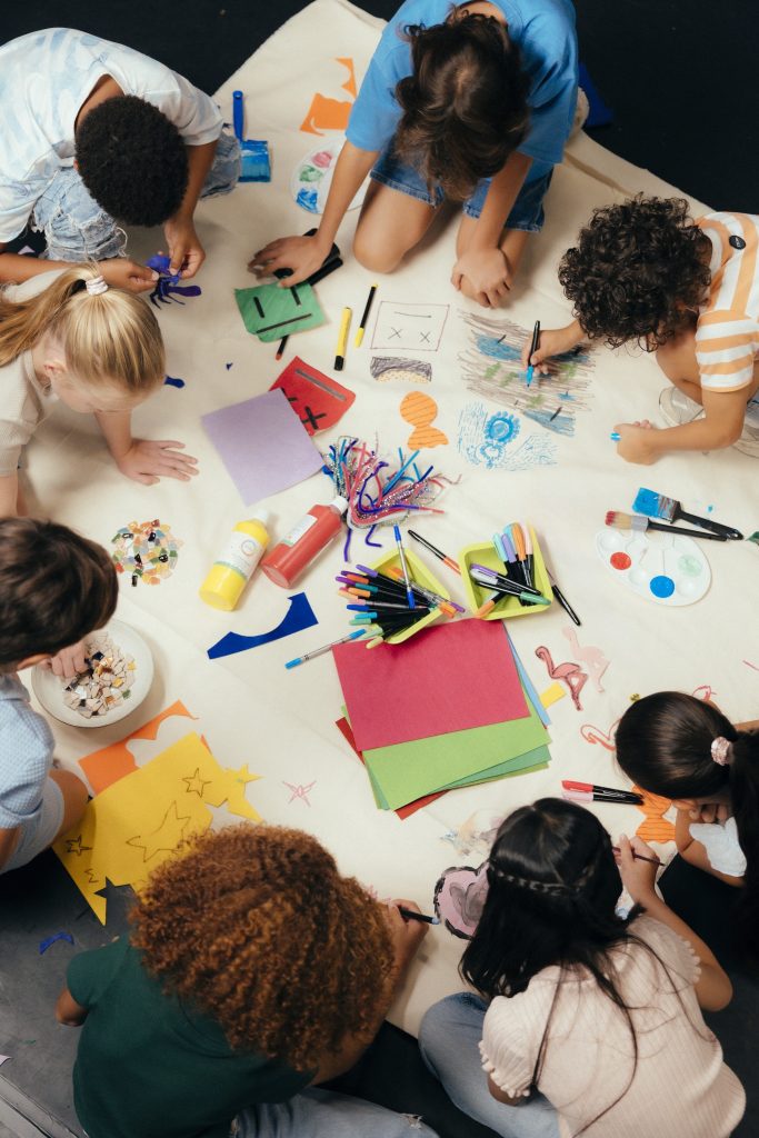 Acht kinderen worden van bovenaf gefotografeerd. Je ziet hun gezichten niet. Ze zitten op een witdoek en zijn aan het knutselen. Op het doek liggen stiften, potloden en verschillende kleuren papier. Het is een vrolijke bedoening.