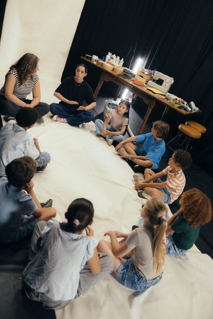 Je ziet kinderen in een kringetje zitten. Ze zitten op het creatieve doek. De 8 kinderen krijgen uitleg van een docent over de creatieve lessen.